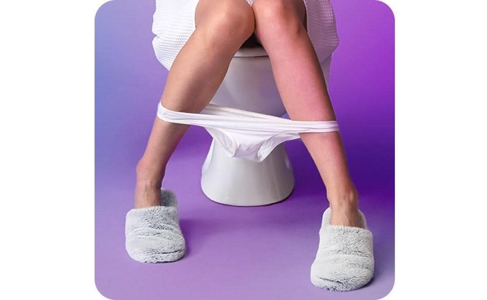 Cropped photo of a woman in slippers sitting on a toilet with her underwear hanging between her legs